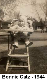 Black and white photo of a baby wearing a knit cap, knit sweater and knee-length dress sitting in a highchair outdoors; houses and leafless trees are visible behind her.